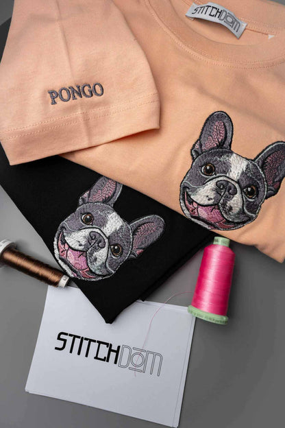 Două tricouri din bumbac organic prezentate într-un aranjament flat-lay: un tricou piersică cu textul „PONGO” brodat pe piept și un tricou negru cu un chip mare de bulldog francez brodat. În jurul lor sunt așezate conuri de ață și un card „Stitchdom” pe o suprafață gri.