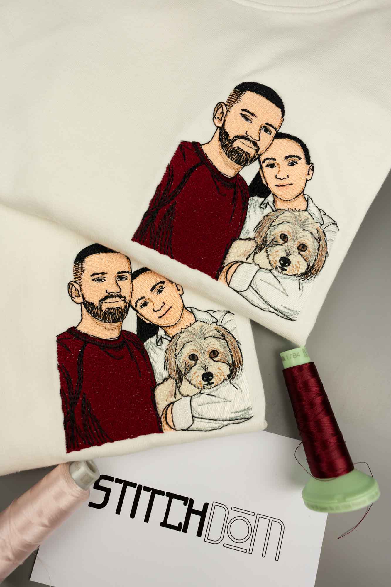 Tricou alb din bumbac organic, cu o poză brodată personalizată cu un cuplu, două persoane și un câine, alături de un con de ață roșu închis și un card cu logo-ul „Stitchdom”, pe un fundal gri neutru.