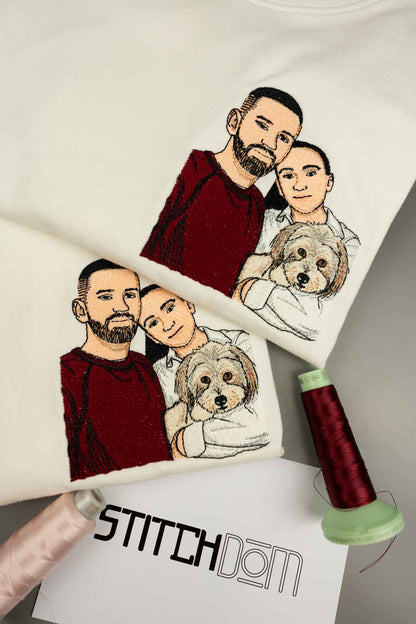 Tricou alb din bumbac organic, cu o poză brodată personalizată cu un cuplu, două persoane și un câine, alături de un con de ață roșu închis și un card cu logo-ul „Stitchdom”, pe un fundal gri neutru.