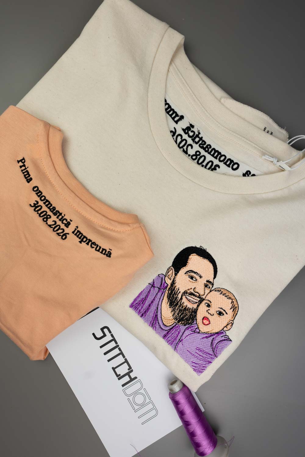 Tricou crem din bumbac organic, împăturit, cu o poză brodată personalizată în stil desen cu un portret de familie și text negru, alături de un tricou piersică, un card alb „Stitchdom'” și un con de ață mov, pe un fundal gri.