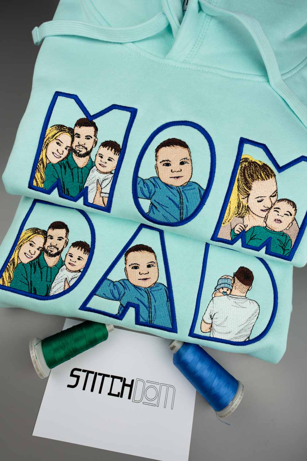 Hanorac verde mentă din bumbac organic, așezat pe o suprafață gri, cu literele mari „MOM DAD” brodate cu contur albastru și umplute cu poze brodate de familie, alături de conuri de ață verde, albă și albastră și un card cu textul „STITCHDOM”.