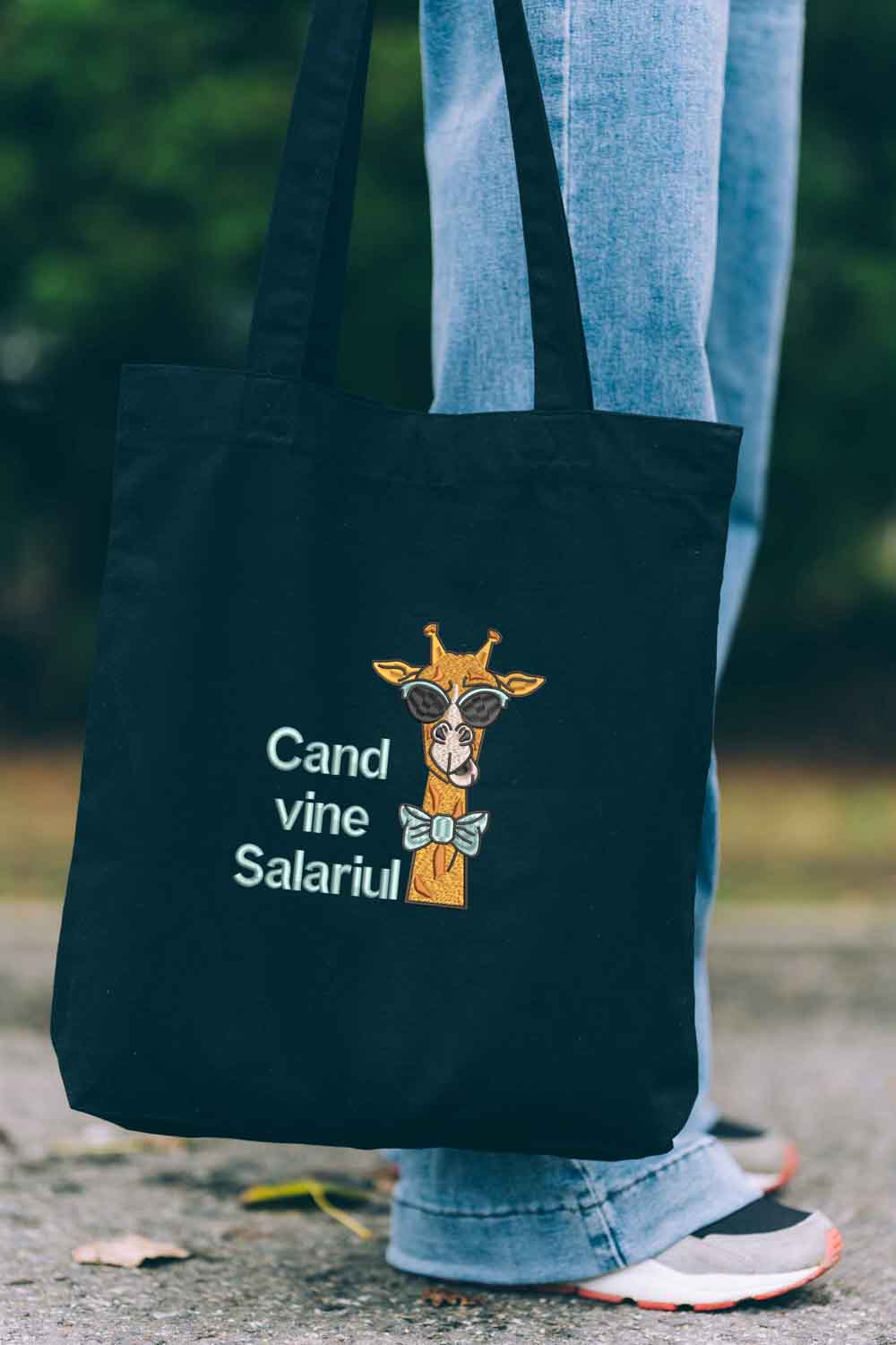 Tote bag brodat negru cu ilustrație amuzantă de girafă purtând ochelari de soare și papion, alături de mesajul „Când vine salariul”, purtat în aer liber.
