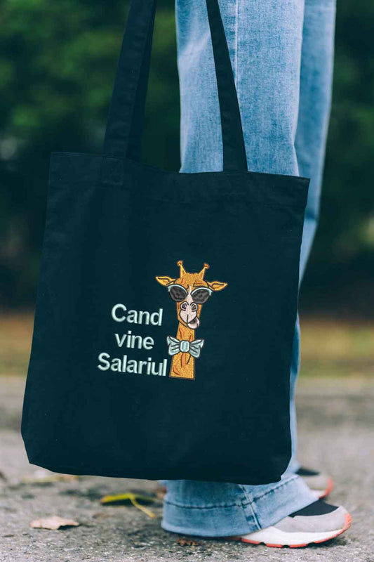 Tote bag brodat negru cu ilustrație amuzantă de girafă purtând ochelari de soare și papion, alături de mesajul „Când vine salariul”, purtat în aer liber.