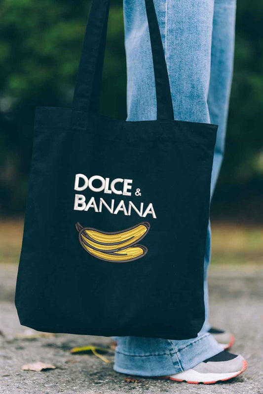 Tote bag brodat negru cu ilustrație de banană și mesajul amuzant „Dolce & Banana”, purtat în aer liber.