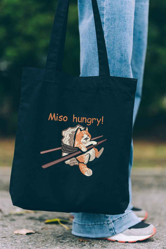 Tote bag brodat negru cu ilustrație amuzantă a unei pisici care mănâncă miso și mesajul „Miso Hungry!”, purtat în aer liber.