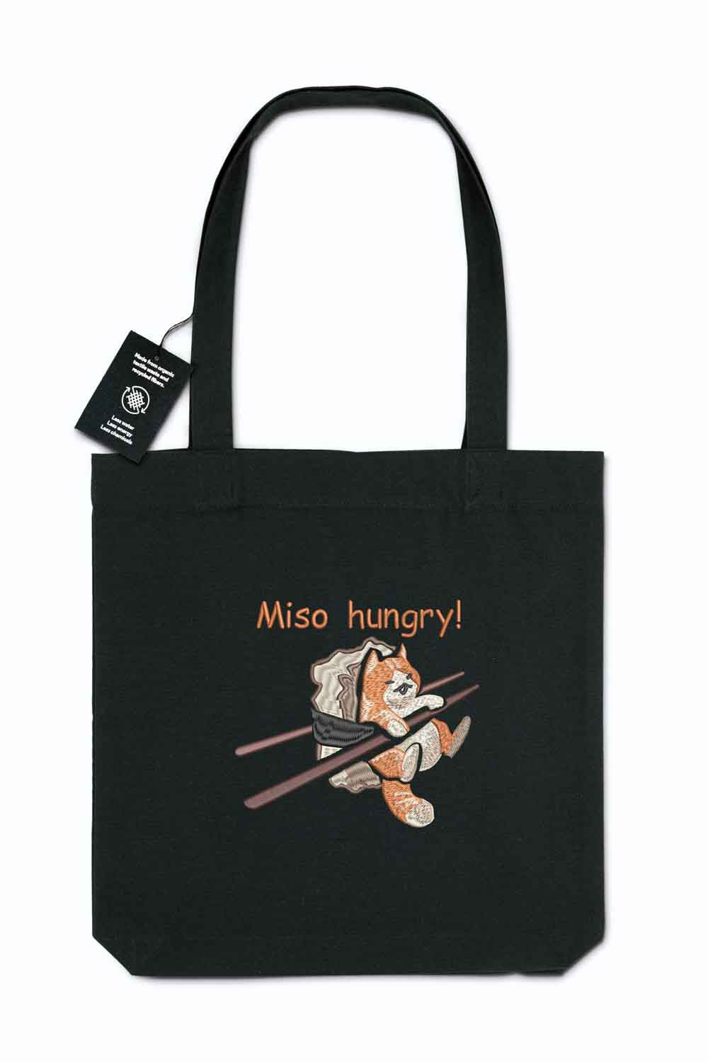 Tote bag brodat negru cu ilustrație amuzantă a unei pisici care mănâncă miso și mesajul „Miso Hungry!”, purtat în aer liber.