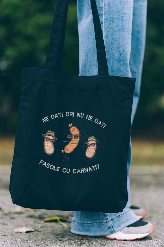 Tote bag brodat negru cu ilustrație amuzantă cu boabe de fasole purtând pălării și ochelari de soare și mesajul românesc „Ne dați ori nu ne dați fasole cu cârnați?”, purtat în aer liber.