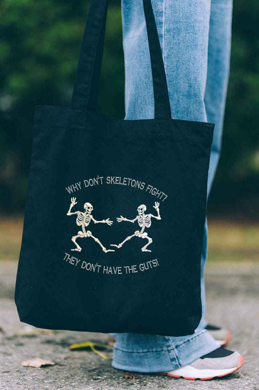 Tote bag brodat negru cu ilustrație albă a două schelete care dansează și mesajul amuzant „Why don't skeletons fight? They don't have the guts!”, purtat în aer liber.
