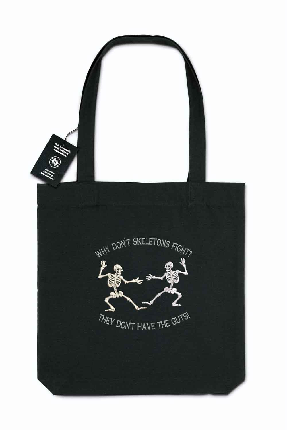 Tote bag brodat negru cu ilustrație albă a două schelete care dansează și mesajul amuzant „Why don't skeletons fight? They don't have the guts!”, purtat în aer liber.