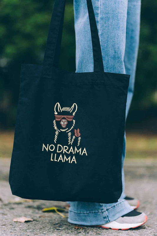 Tote bag brodat negru cu ilustrație de lamă purtând ochelari de soare și mesajul amuzant „No Drama Llama”, purtat în aer liber.