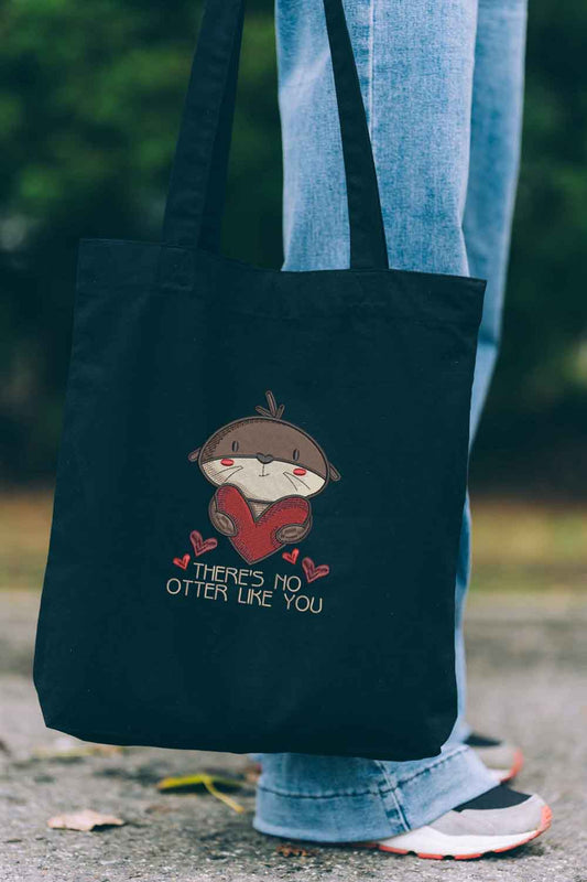 Tote bag brodat negru cu ilustrație drăguță de vidră care ține o inimă și mesajul amuzant „There's No Otter Like You”, purtat în aer liber.