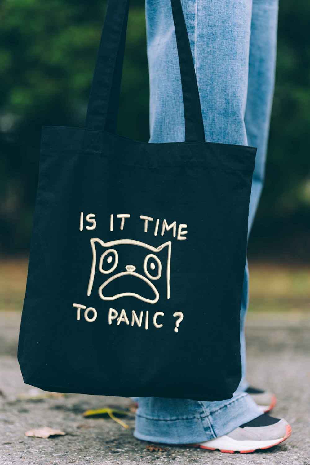 Tote bag brodat negru cu ilustrație amuzantă a unei pisici îngrijorate și mesajul „Is It Time To Panic?”, purtat în aer liber.