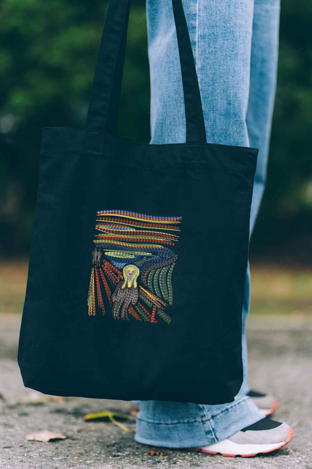 Tote bag brodat negru cu design colorat reprezentând un dinte înconjurat de pene, inspirat de „The Scream”, purtat în aer liber.