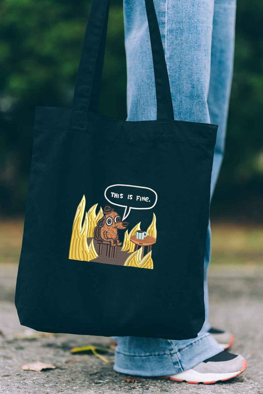 Tote bag brodat negru cu ilustrație amuzantă a unui câine în flăcări și mesajul „This Is Fine”, purtat în aer liber.