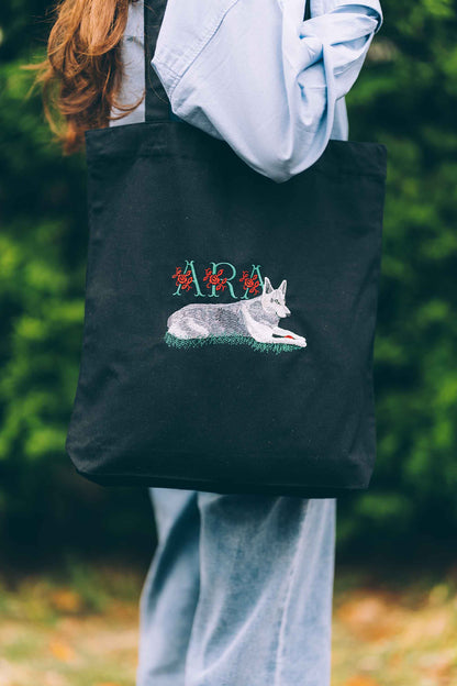 Geantă tote neagră din bumbac organic, ținută de o persoană, cu o poză brodată cu un câine și textul „ARA”, într-un cadru exterior.