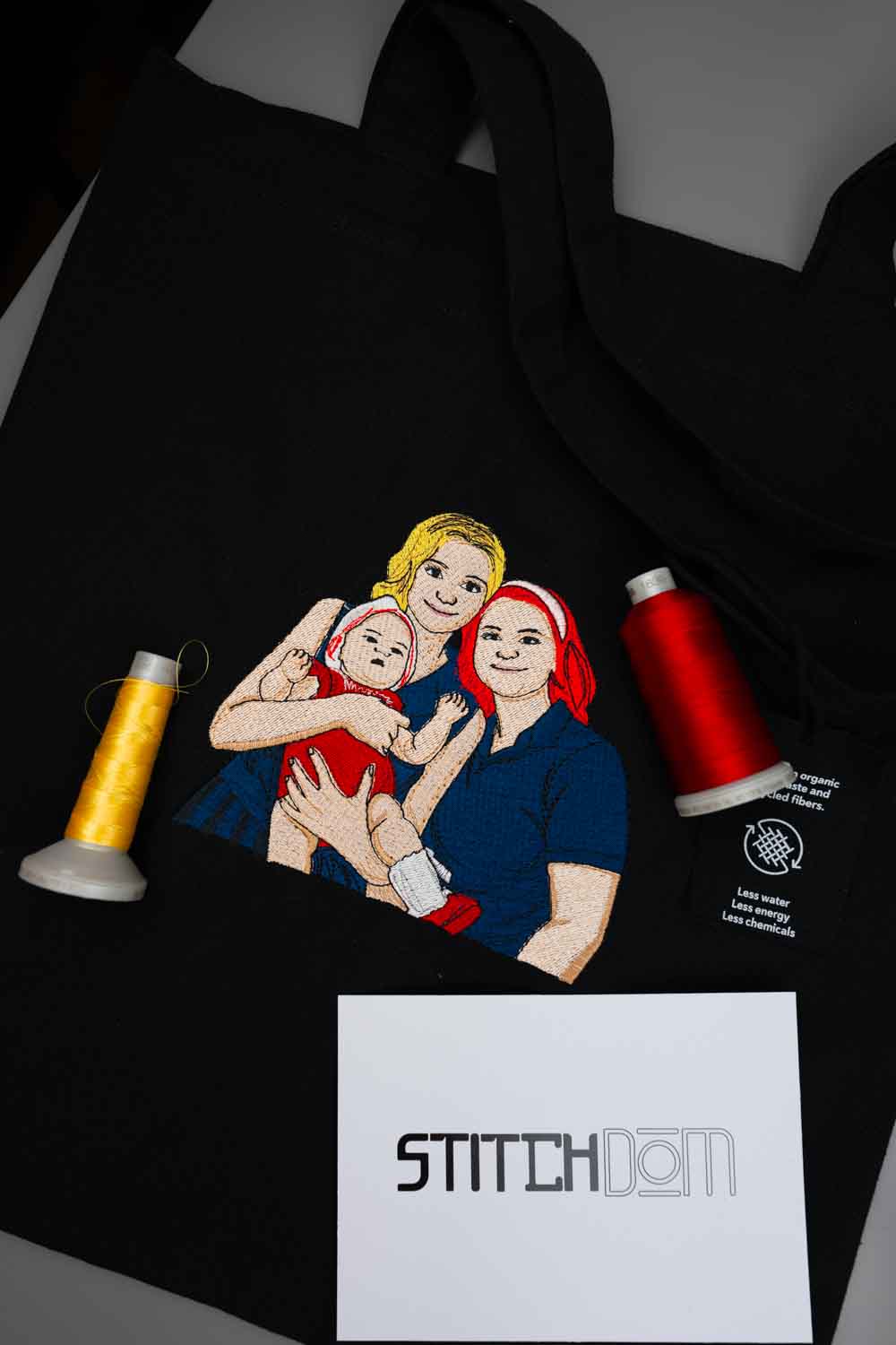 Geantă tote neagră din bumbac organic, așezată flat lay pe o suprafață gri, cu o poză brodată colorată cu o mamă care își ține bebelușul în brațe, alături de conuri de ață și un card alb cu logo-ul „Stitchdom”.