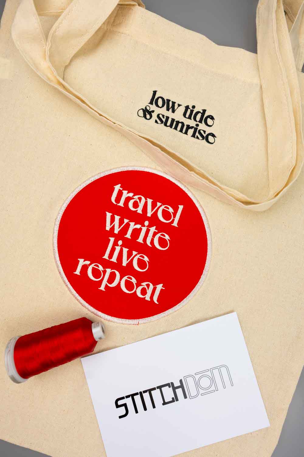 Geantă tote bej din pânză, cu textul negru „low tide & sunrise”, alături de un sticker circular roșu cu mesajul „travel write live repeat”, un con de ață roșie și un card cu logo-ul „Stitchdom”, pe un fundal neutru.