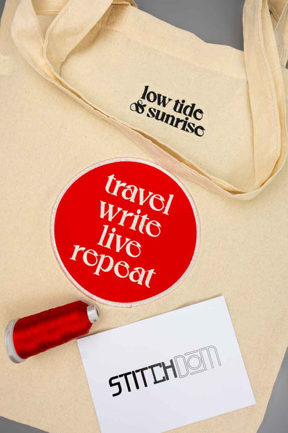 Geantă tote bej din pânză, cu textul negru „low tide & sunrise”, alături de un sticker circular roșu cu mesajul „travel write live repeat”, un con de ață roșie și un card cu logo-ul „Stitchdom”, pe un fundal neutru.